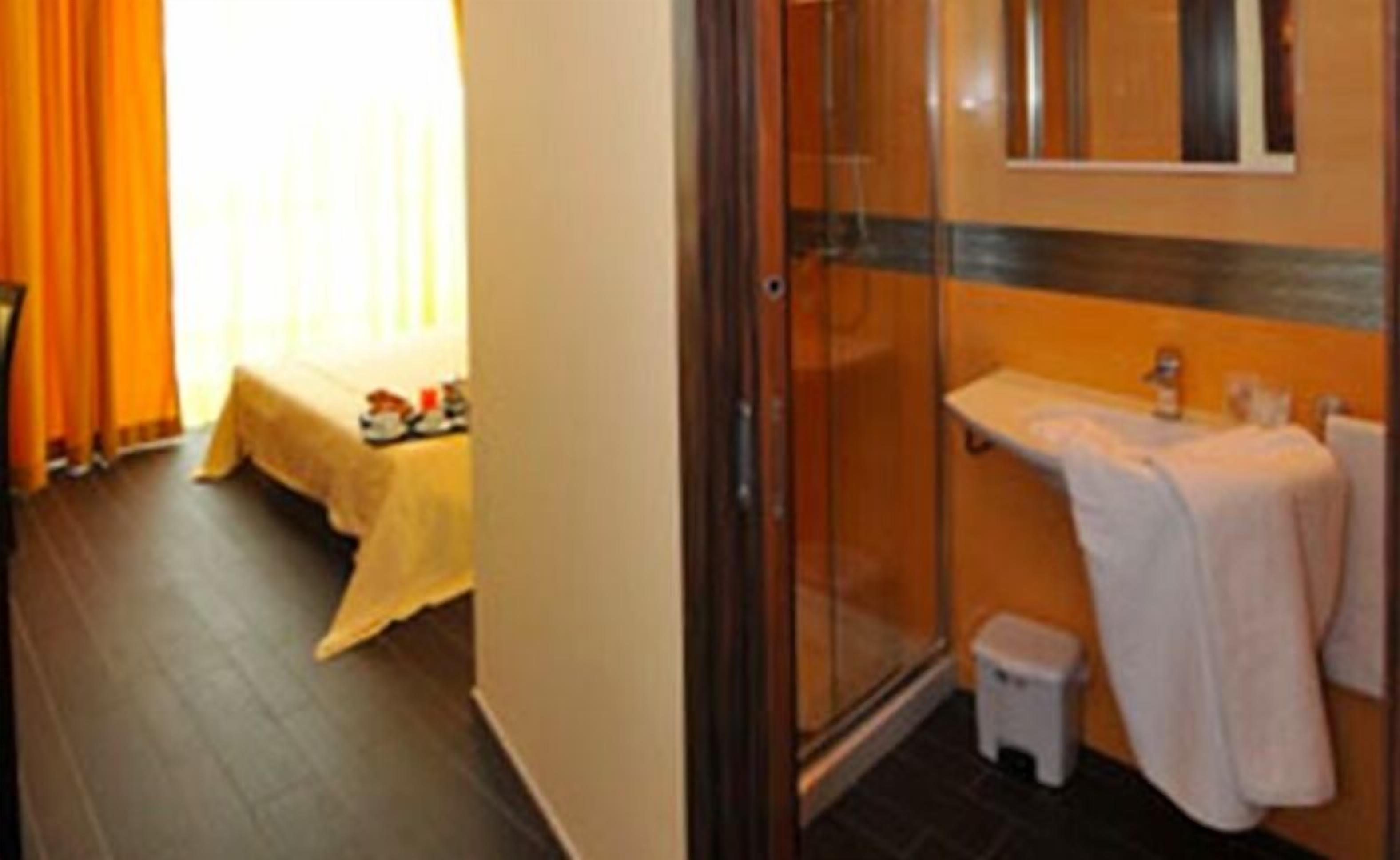 Habitación de hotel con cama, una mesita con objetos, zona de ducha y lavamanos con toalla.