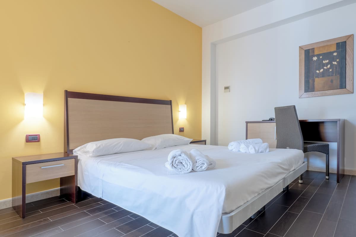 A neatly made bed with white linens and towels, a bedside table with a lamp, a desk with a chair, and a painting on the wall.