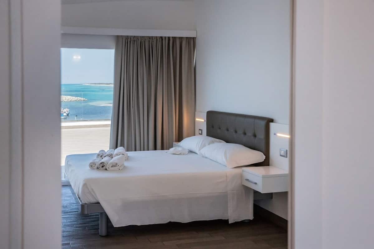 A hotel room with a bed, a nightstand, a window with a view of the sea, and a curtain.