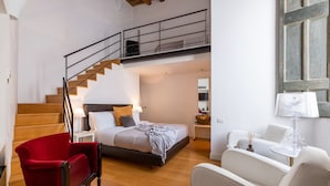 Un dormitorio con cama, una silla roja, un sillón blanco, una mesita y una escalera que lleva a un nivel superior.