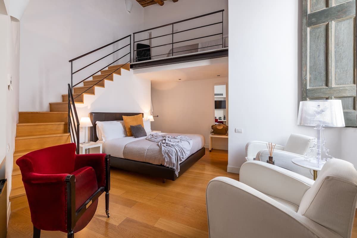 Une chambre à coucher avec un lit, une chaise rouge, un fauteuil blanc, une petite table et un escalier menant à un niveau supérieur.