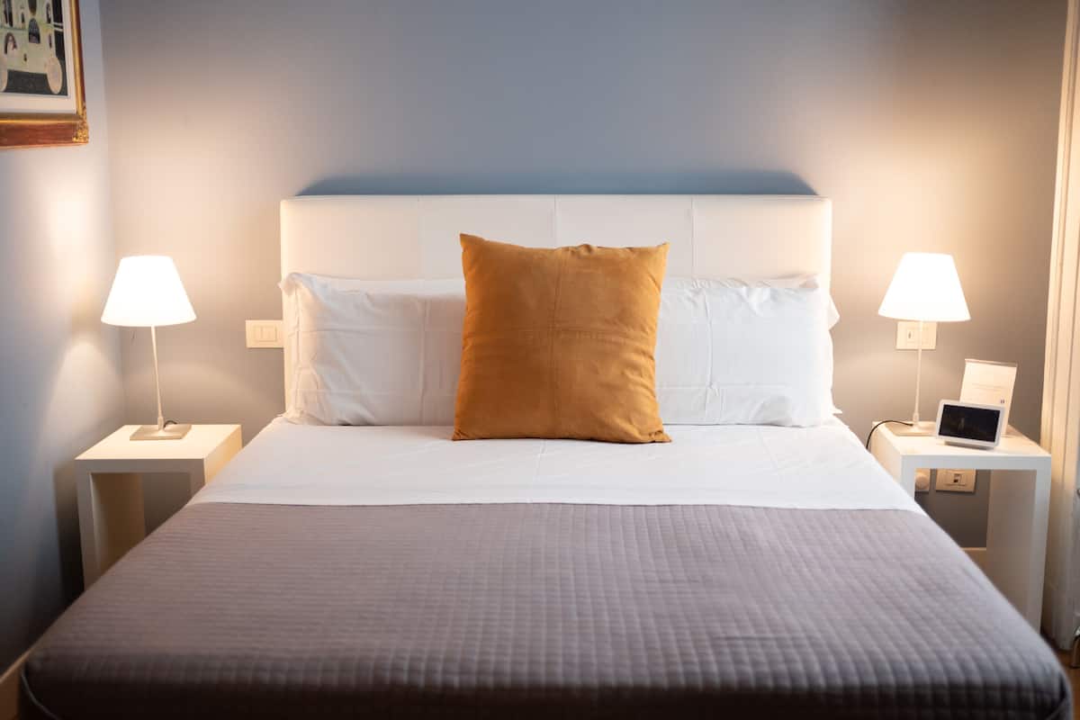 Un lit bien fait, avec du linge de lit blanc et un seul oreiller marron, flanqué de deux tables de chevet sur lesquelles sont posées des lampes.