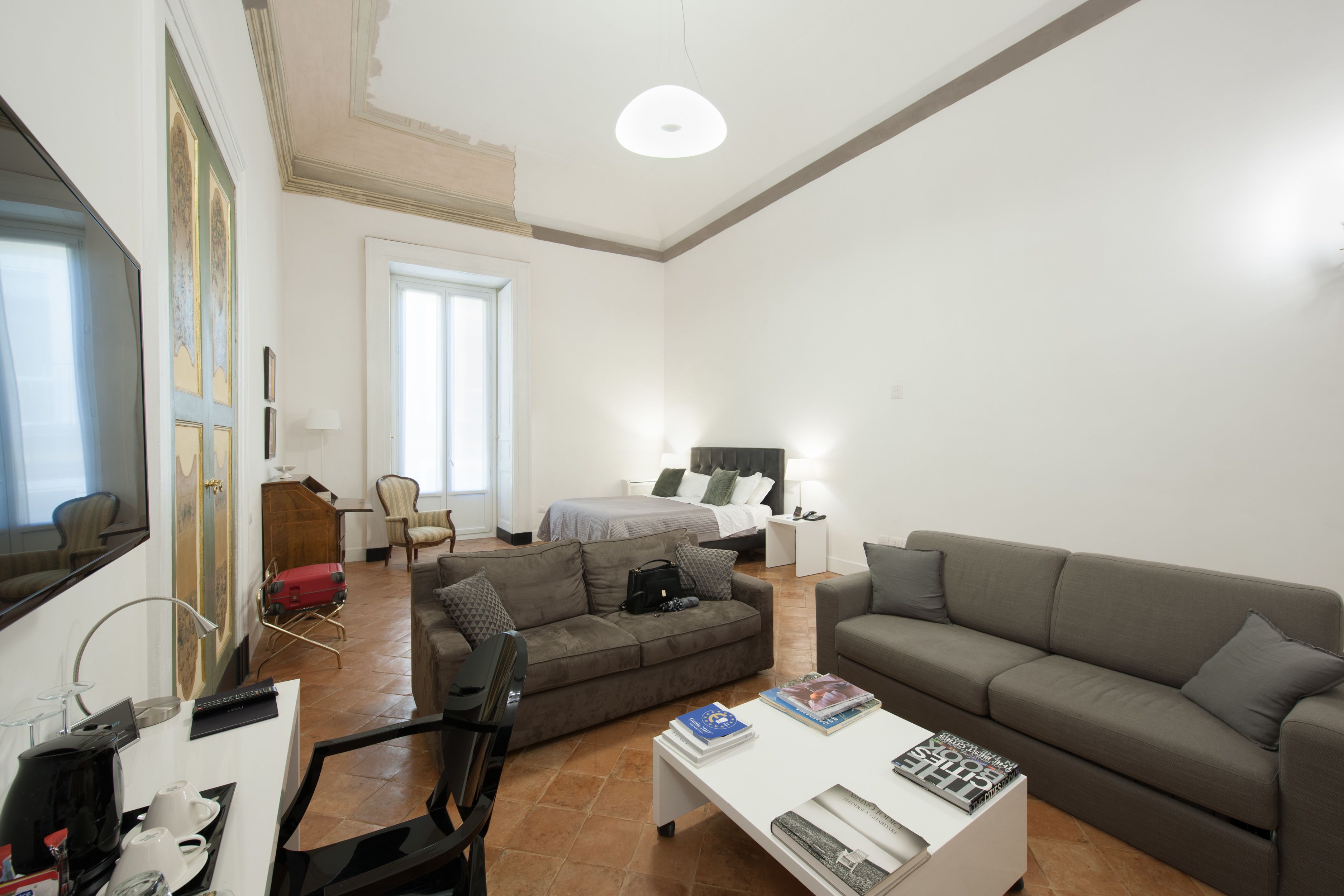 Un salon moderne avec un canapé, un fauteuil, une table basse et un téléviseur. On aperçoit, au fond, un lit avec une table de chevet et une lampe.