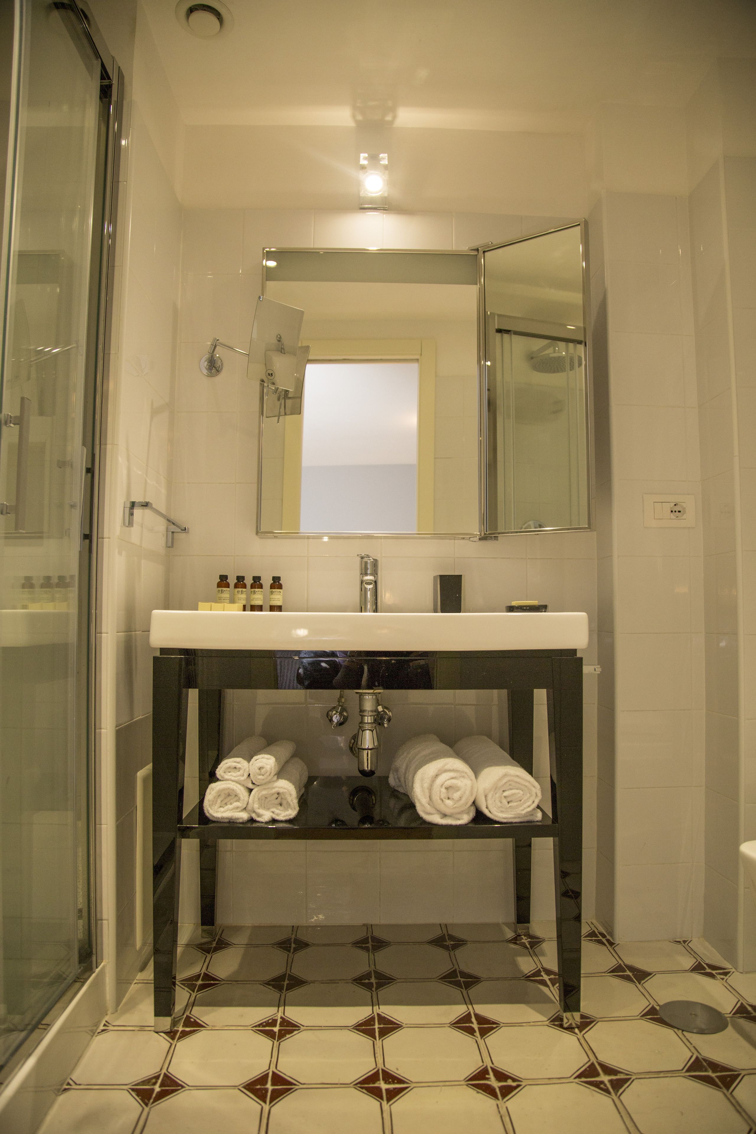 A bathroom with a glass shower enclosure, a white sink with a dark countertop, a mirror above the sink, and a tiled floor with a geometric pattern.