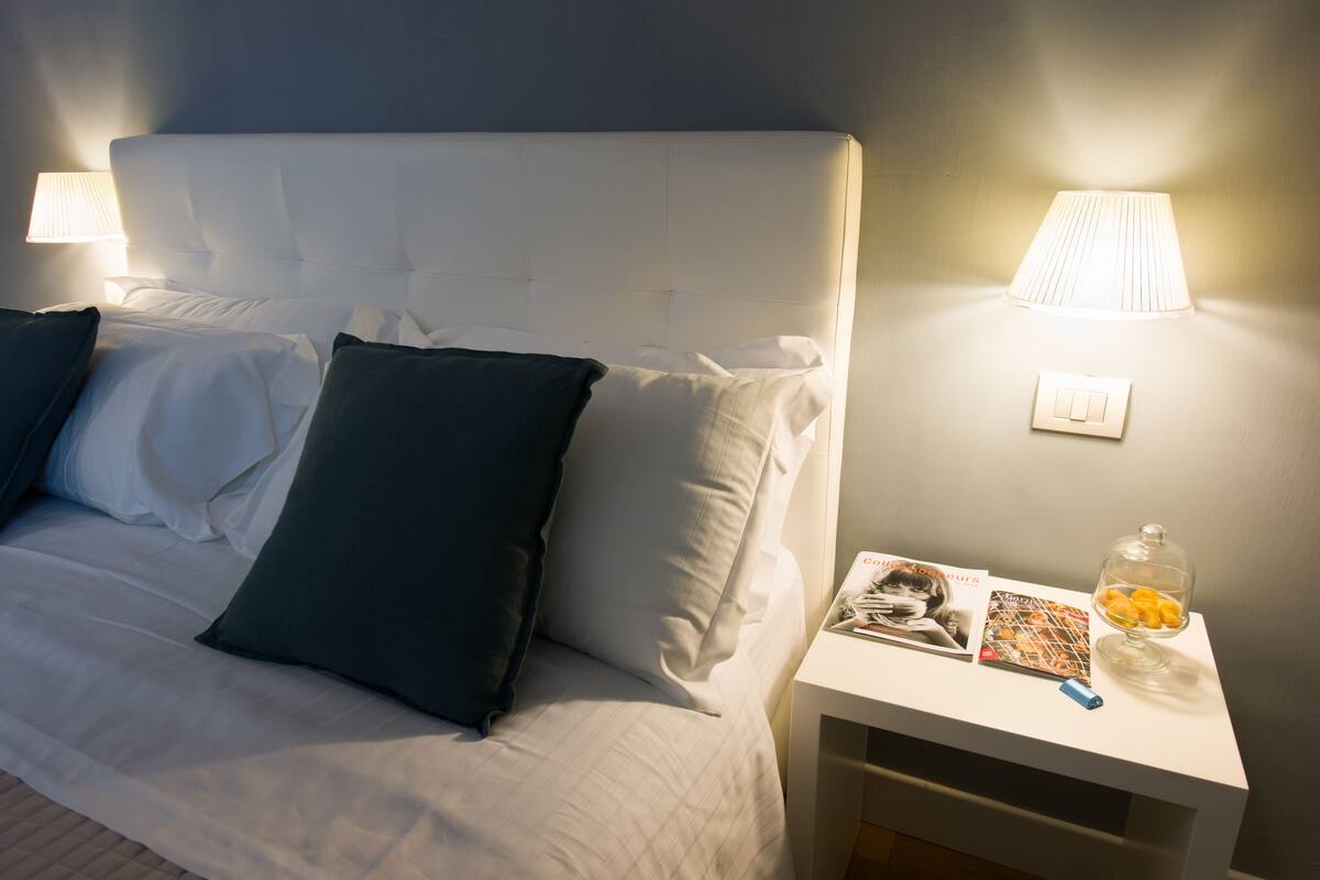 Un lit bien fait, avec des oreillers blancs et bleu foncé, une table de chevet sur laquelle se trouvent une lampe, un livre et un bocal rempli de collations.