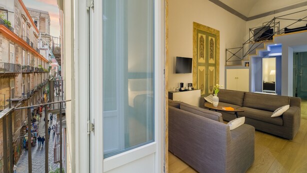 Un apartamento moderno con balcón que da a una calle estrecha, una sala de estar con sofá y una mesa de centro, y una escalera que conduce a un piso superior.
