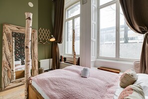 Ein Schlafzimmer mit großem Fenster, einem Bett mit rosa Steppdecke und einem Spiegel mit Holzrahmen.