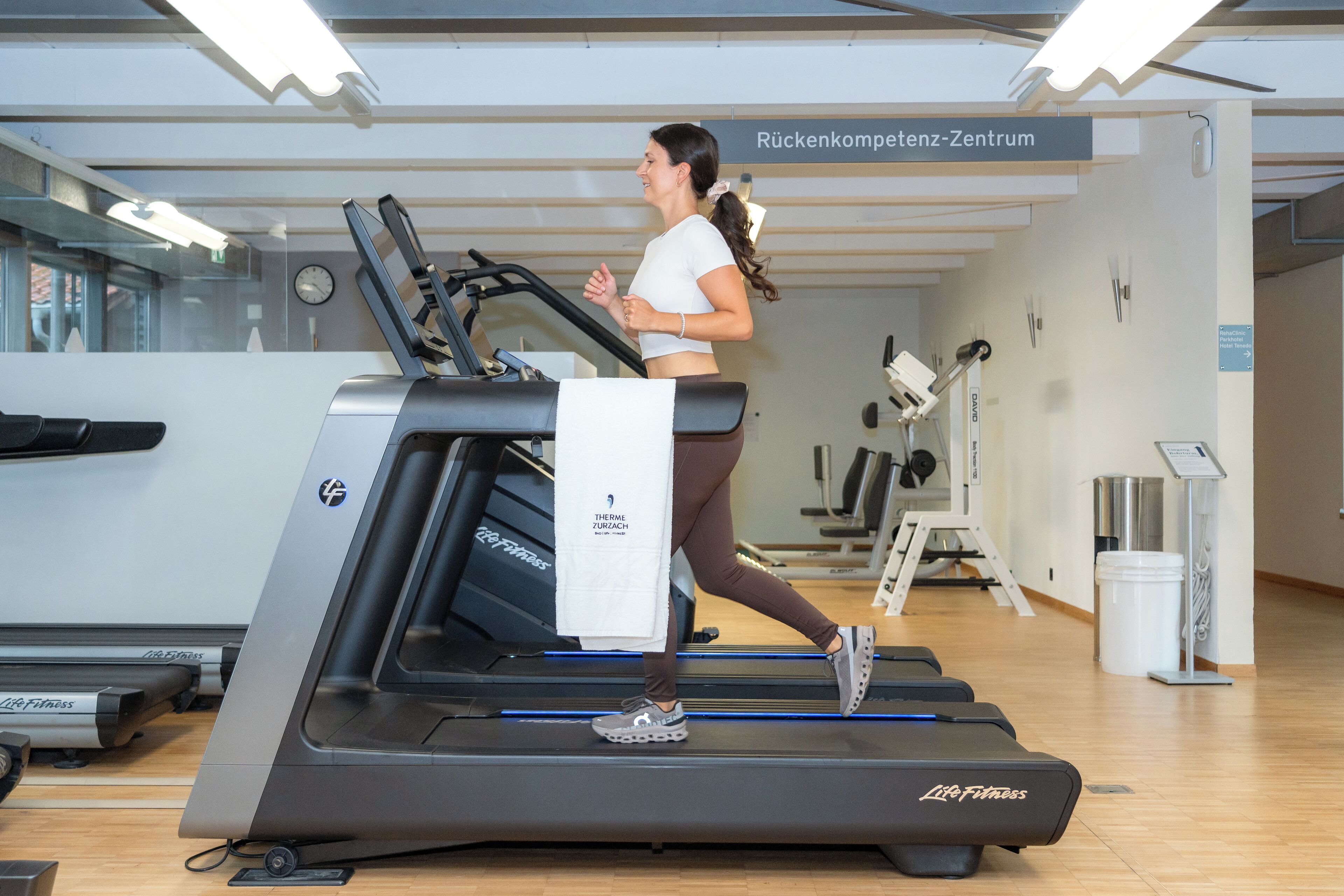 A woman running on a treadmill in a gym with a visible sign reading "Rückenkompens-Zentrum".