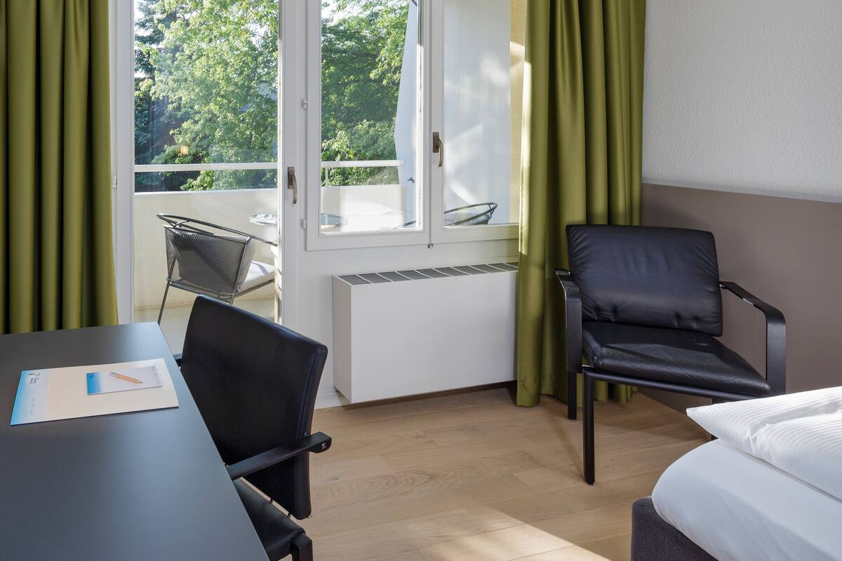 A hotel room with a bed, a desk with a chair, a window with curtains, and a view of trees outside.