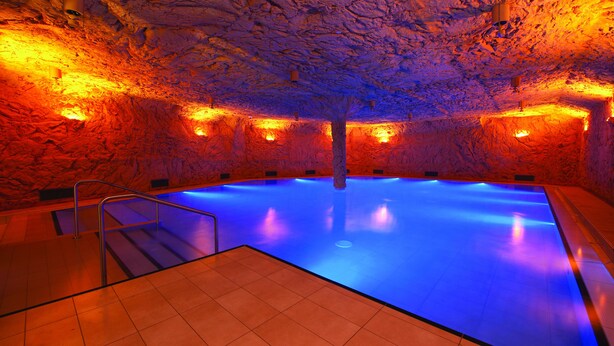 An indoor swimming pool with a blue-lit water feature, surrounded by a textured wall and warm lighting. - VitalBoutique Hotel Zurzacherhof (Bad Zurzach)