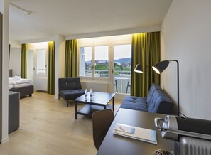 A hotel room with a bed, a sofa, a desk, and a chair. There is a window with curtains, a radiator, and a lamp. - VitalBoutique Hotel Zurzacherhof (Bad Zurzach)