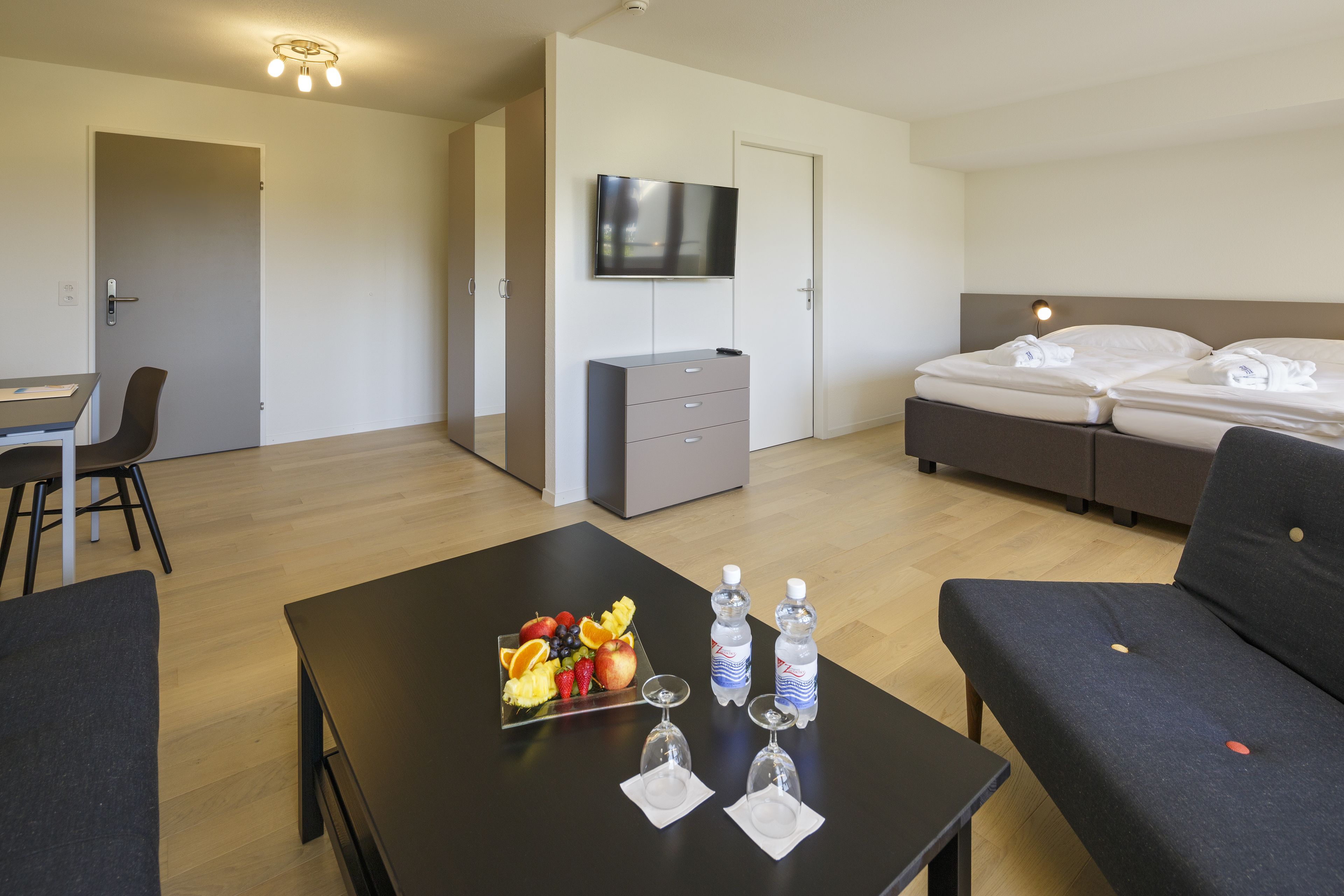 A hotel room with a bed, a TV, a desk with a chair, a small dresser, and a table with fruit and water bottles.