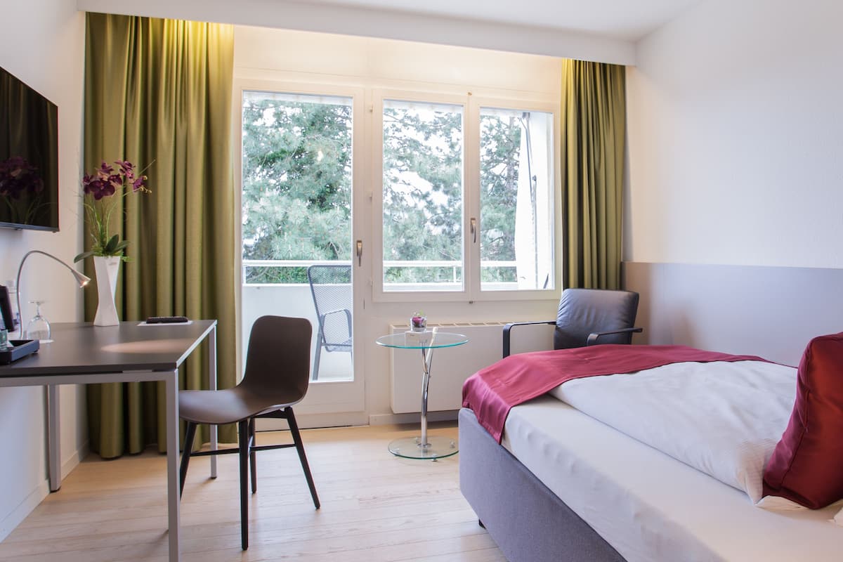 A bedroom with a bed, a desk, a chair, a small table, and a window with curtains.