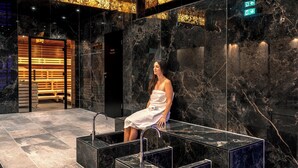 A woman sitting on a black marble bench in a modern lobby with a marble wall and a wooden reception desk in the background. - VitalBoutique Hotel Zurzacherhof (Bad Zurzach)