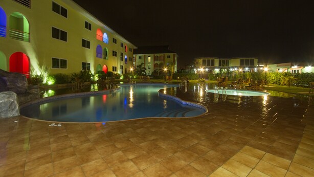 A swimming pool area with tiled flooring, a building with multiple windows and balconies, and a well-lit exterior. - Kingstel Hotel (Sekondi-Takoradi)