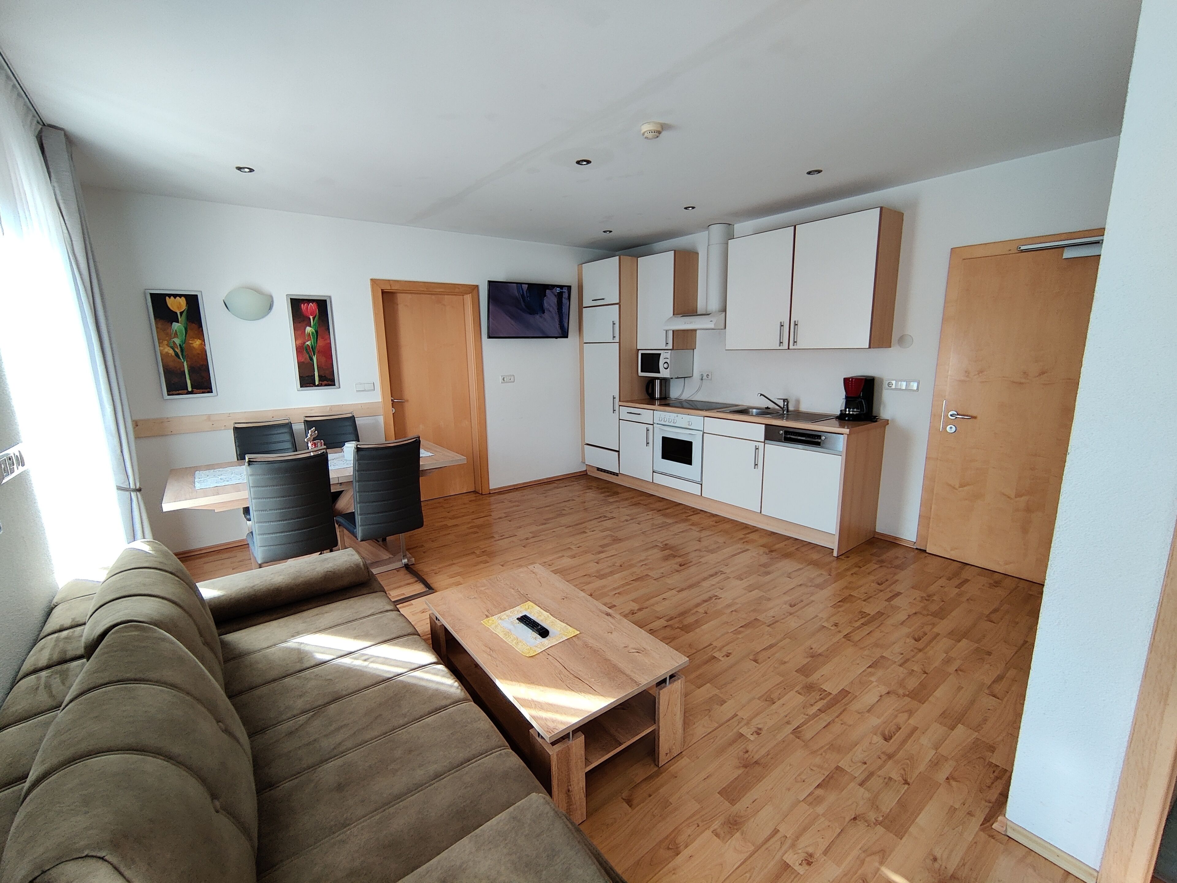 Una sala moderna con sofá y una mesa de centro, un comedor con sillas, una cocina con armarios y electrodomésticos, y una puerta que da a otra habitación.