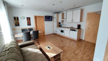 Una sala moderna con sofá y una mesa de centro, un comedor con sillas, una cocina con armarios y electrodomésticos, y una puerta que da a otra habitación.