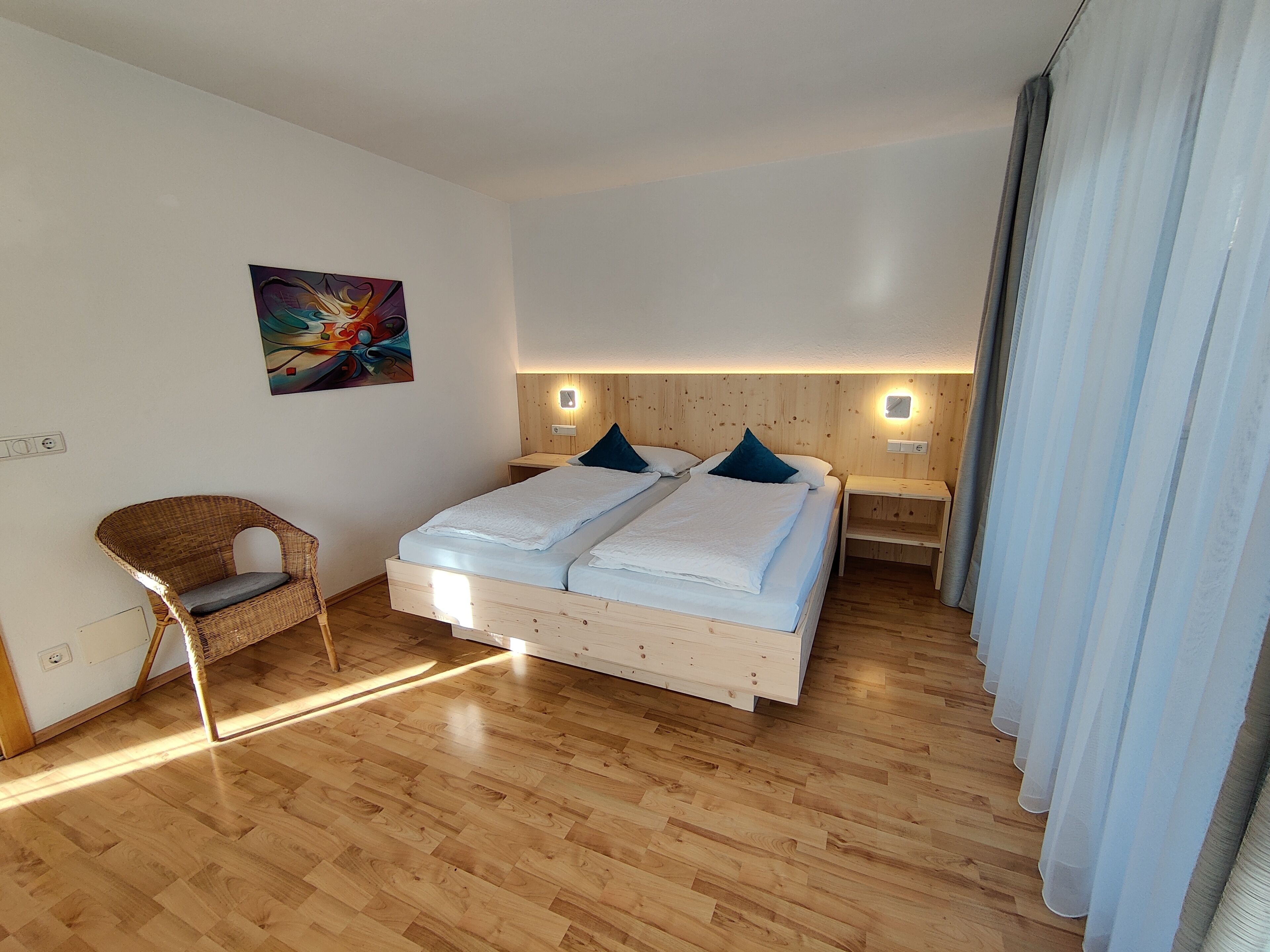 Un dormitorio con piso de madera, una cama con sábanas blancas, dos mesitas de noche, una silla de mimbre y un cuadro en la pared.