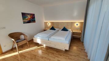 Un dormitorio con piso de madera, una cama con sábanas blancas, dos mesitas de noche, una silla de mimbre y un cuadro en la pared.