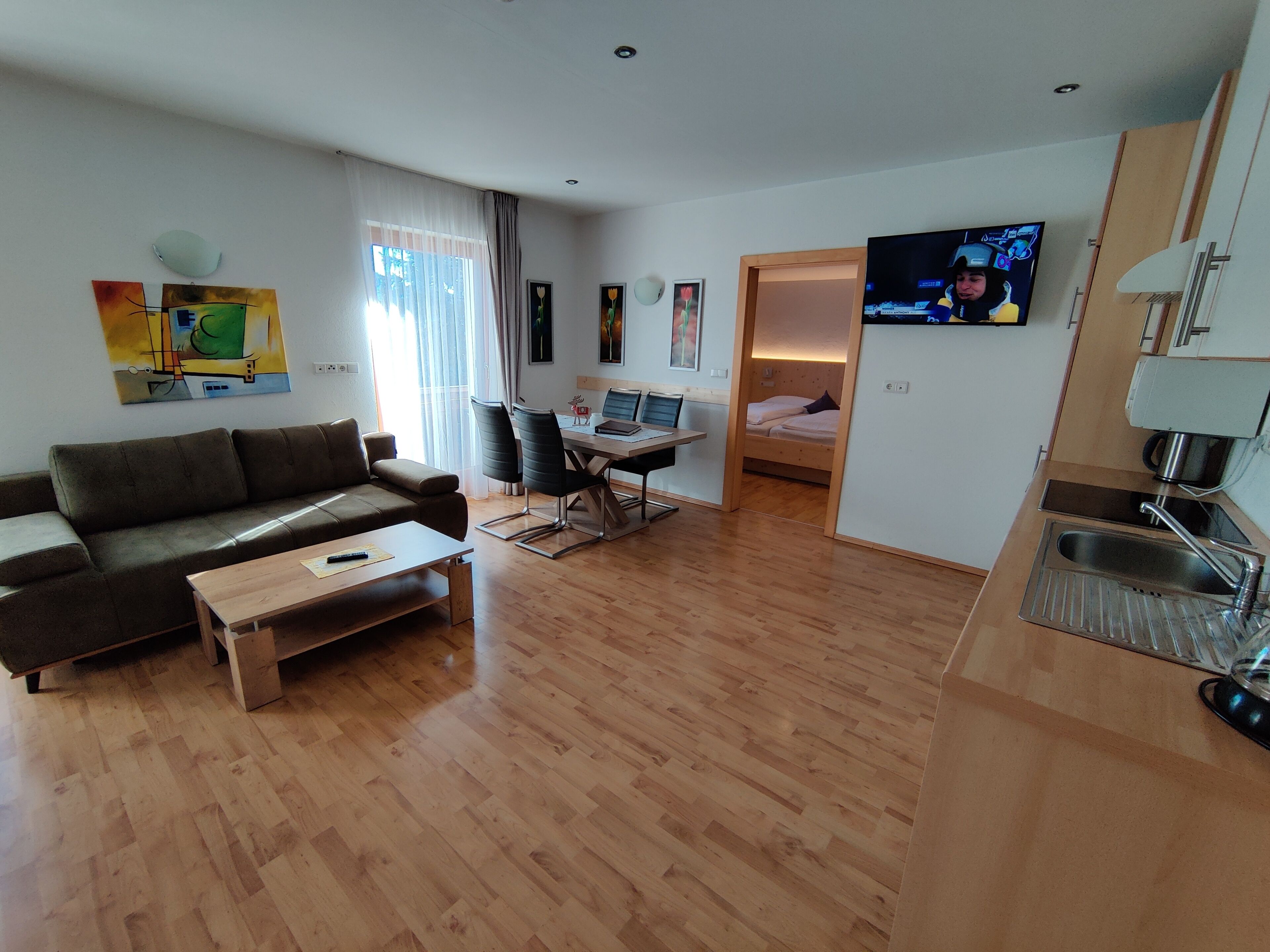 A modern living room with a sofa, coffee table, and a TV. There is a dining area with a table and chairs, and a kitchen with a sink and stove.