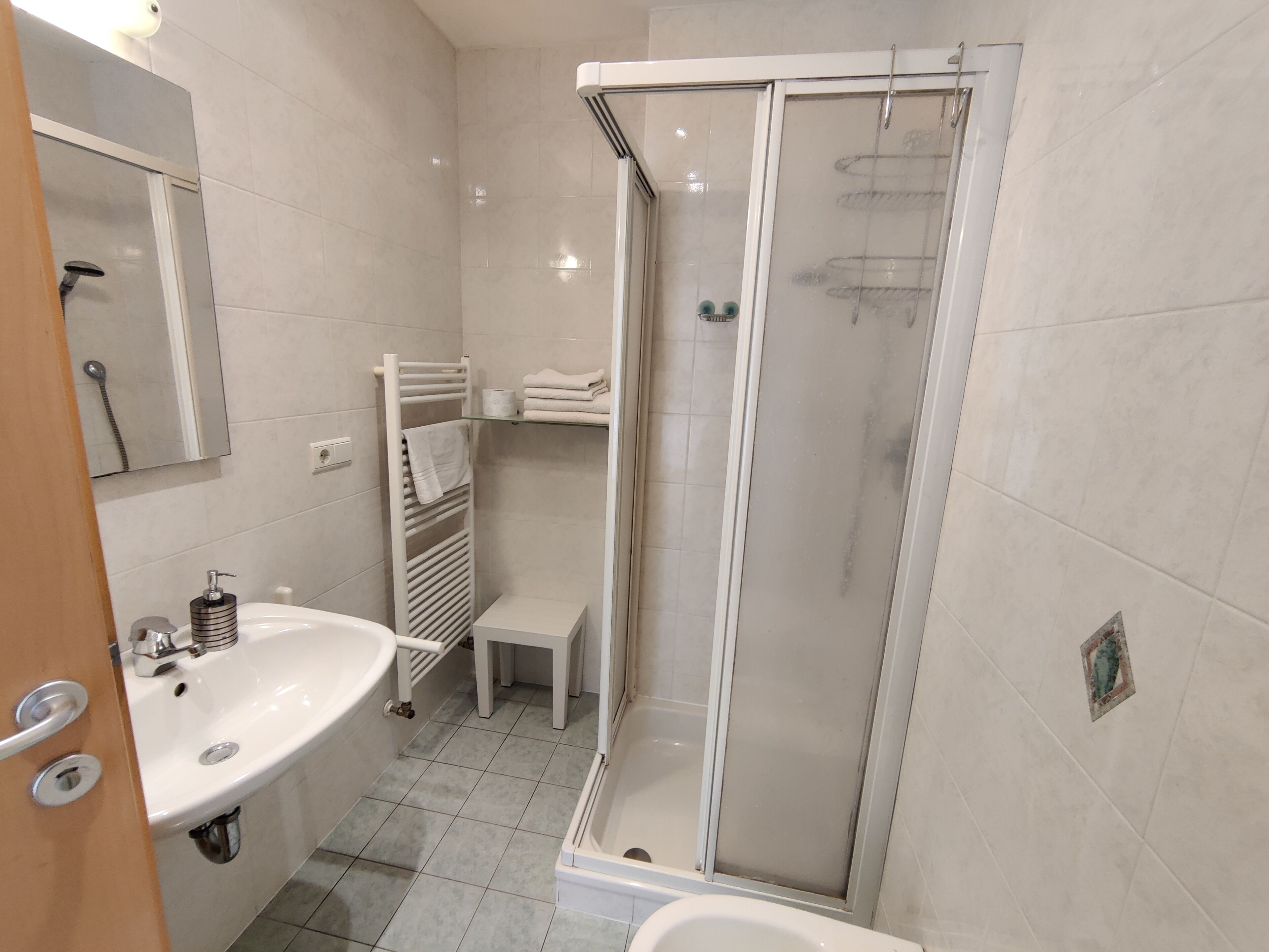 Un bany modern amb dutxa d'ús de vidre, un lavabo blanc i un petit tamboret.