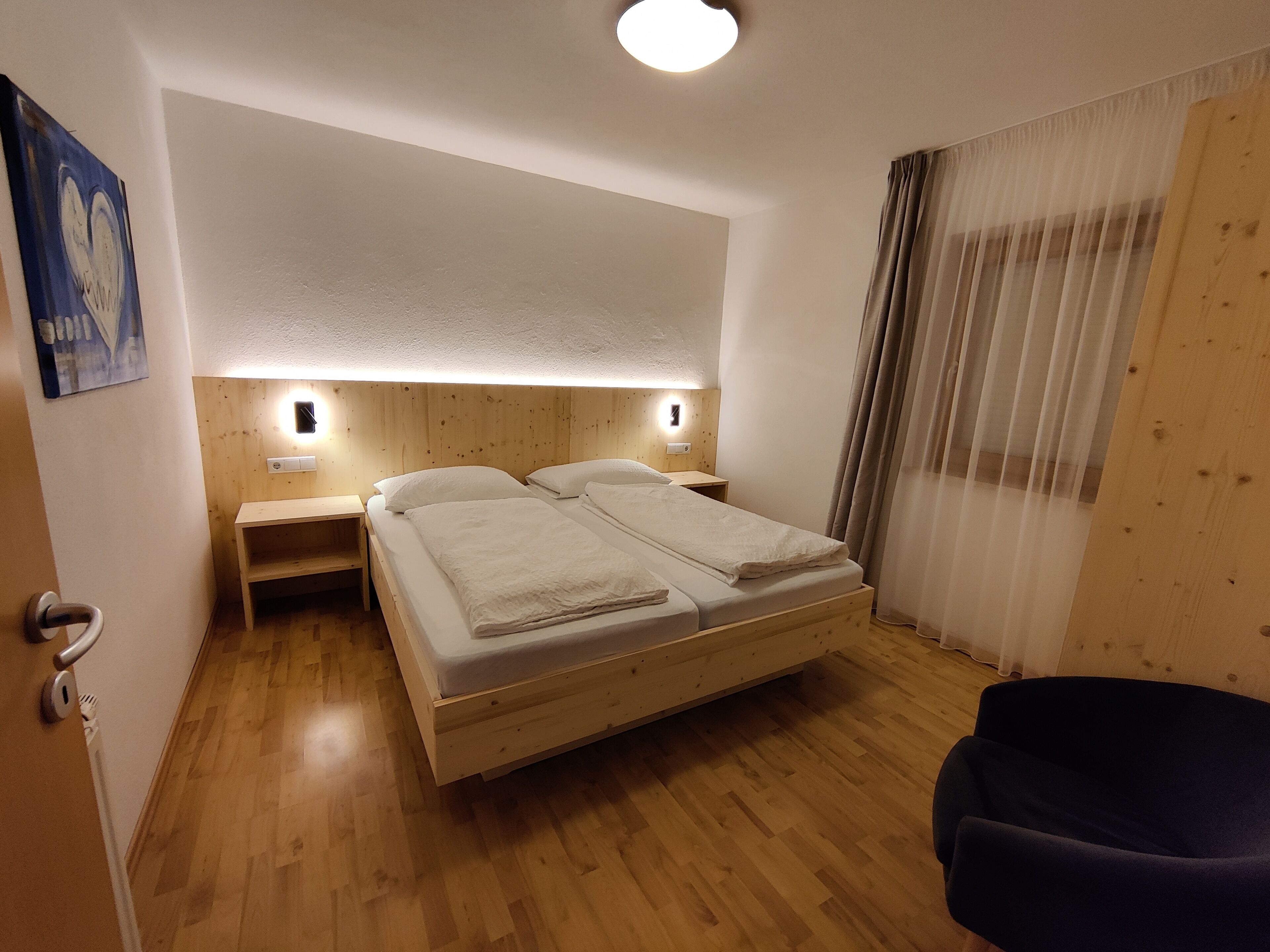 A hotel room with a wooden bed, bedside tables, a nightstand, a chair, and a painting on the wall.