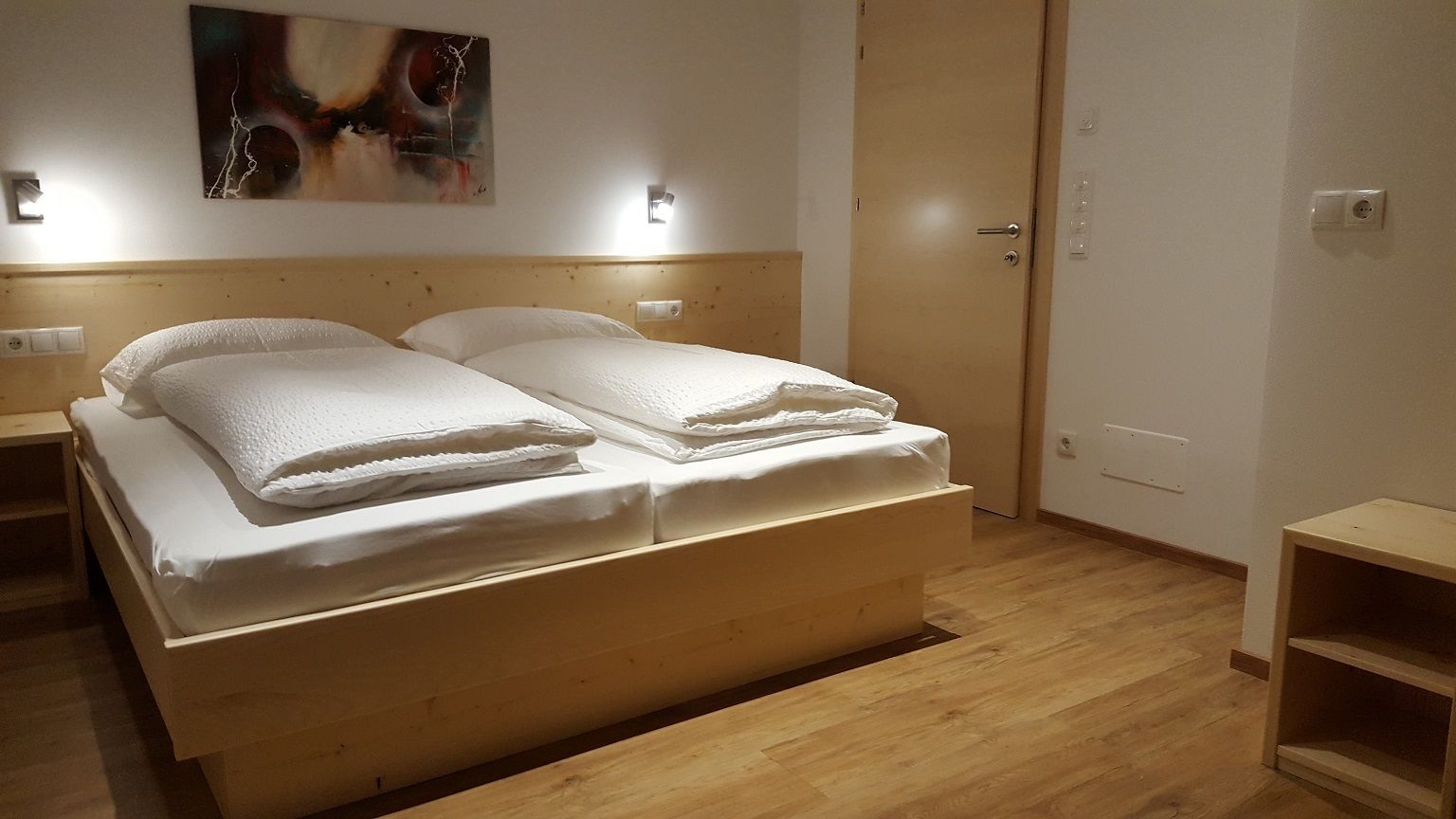 A double bed with white linens, a wooden headboard, a wall-mounted light, and a door to the right.