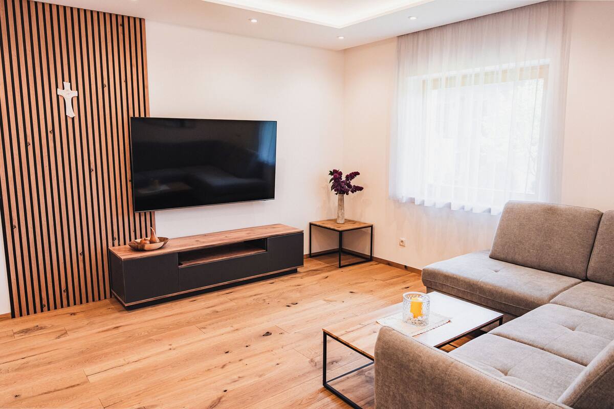 Una sala de estar moderna con un televisor de pantalla plana, piso de madera, un sofá gris y una mesita auxiliar con un jarrón.