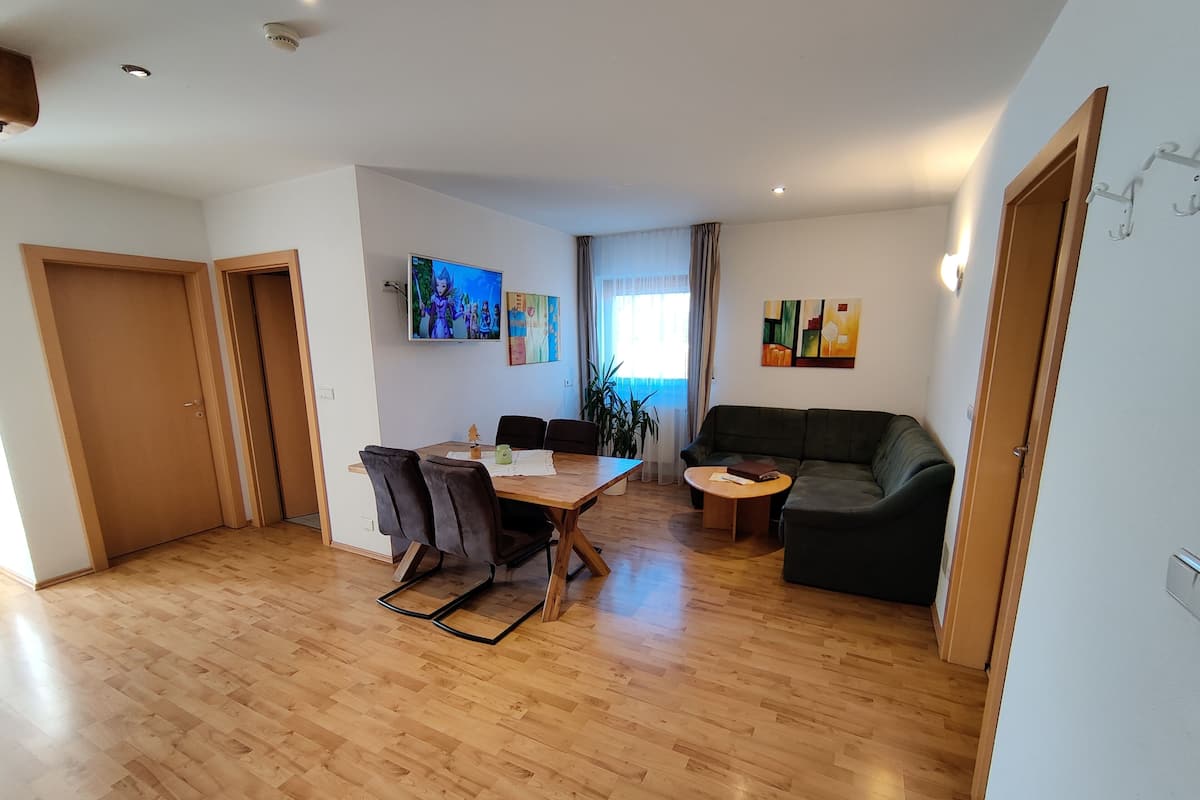 Un salón espacioso con suelo de madera, una mesa de comedor con sillas, un sofá y un televisor montado en la pared.
