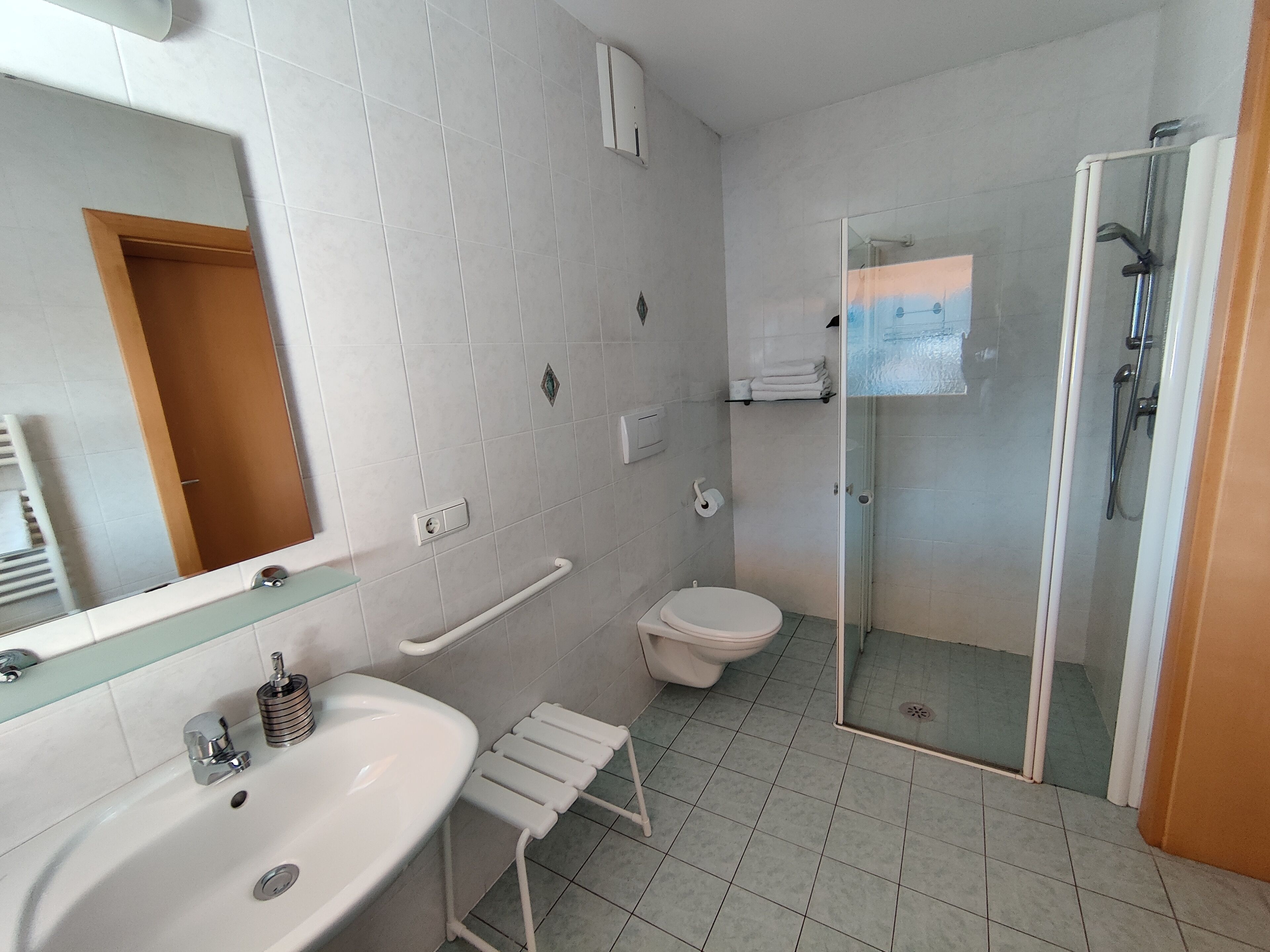 Une salle de bain moderne avec un lavabo, un miroir et des toilettes blancs. Elle comprend une douche en verre et une barre d’appui pour faciliter l’accès aux personnes à mobilité réduite.