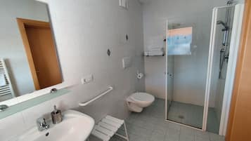 Une salle de bain moderne avec un lavabo, un miroir et des toilettes blancs. Elle comprend une douche en verre et une barre d’appui pour faciliter l’accès aux personnes à mobilité réduite.