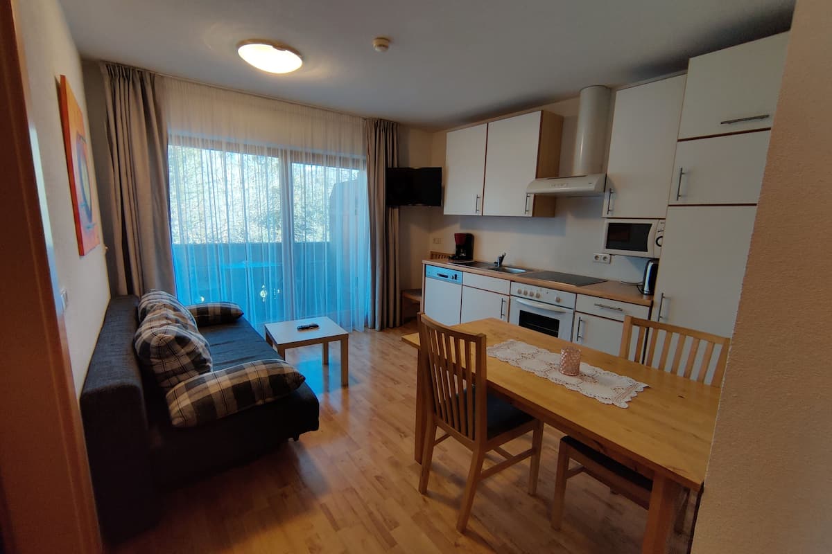 Un espacio habitable compacto con sofá, mesa de comedor con sillas, zona de cocina con electrodomésticos y un amplio ventanal con cortinas.
