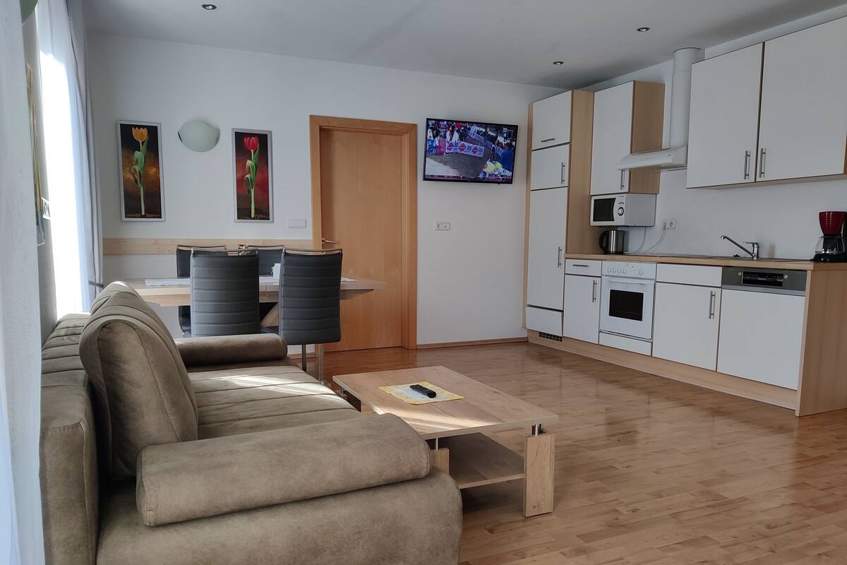 Una sala moderna con sofá, una mesa de centro y un comedor con sillas. Hay una cocina con armarios y electrodomésticos blancos.
