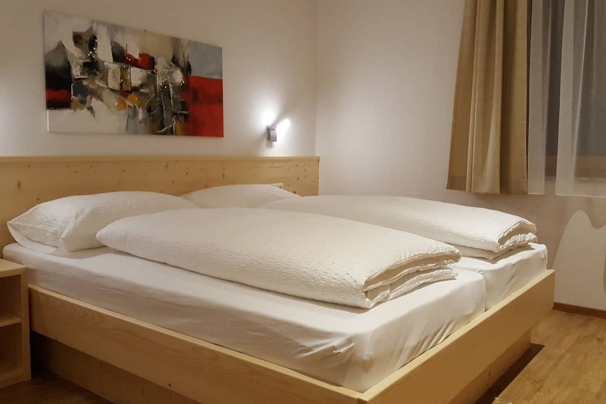Cama doble con sábanas blancas en una habitación con cabecera de madera, un cuadro abstracto colgado en la pared y una ventana con cortinas translúcidas.
