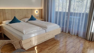 Uma cama de madeira com duas almofadas, um criado-mudo, uma cadeira de vime e uma janela com cortinas leves.