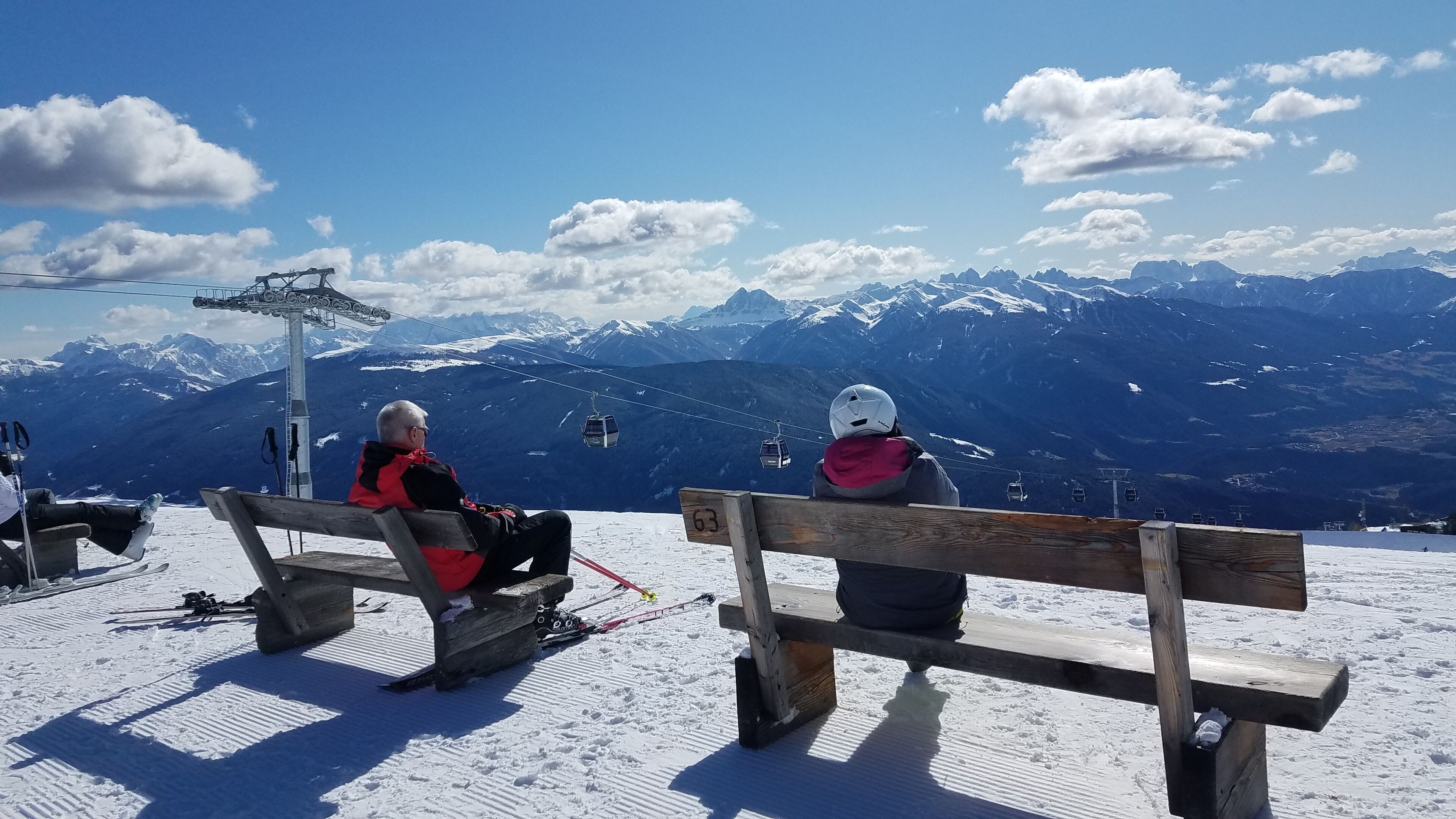 Zwei Personen sitzen auf hölzernen Bänken und blicken auf eine verschneite Berglandschaft mit Sesselliften im Hintergrund.