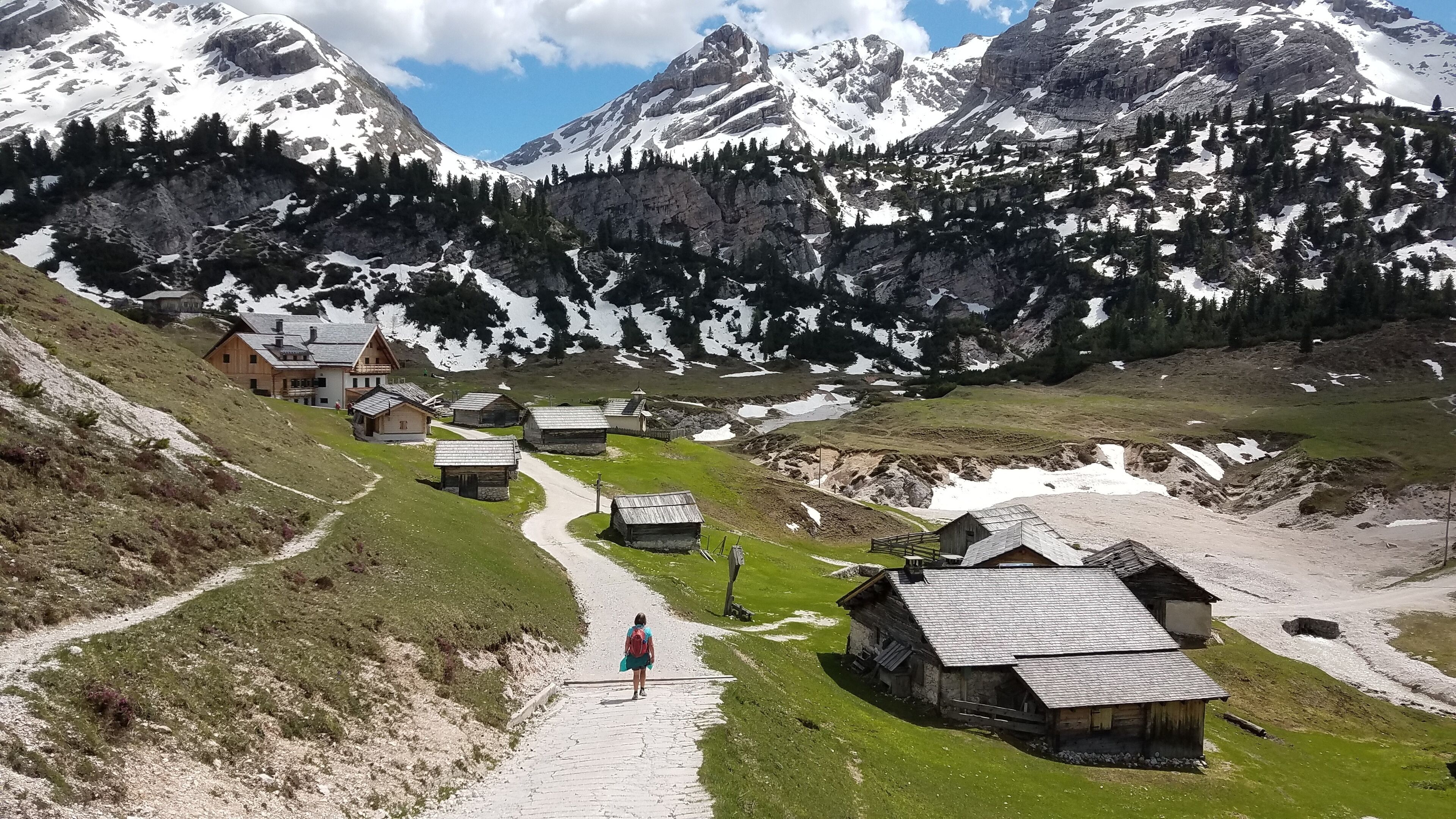 Ein Bergdorf mit traditionellen Holzhäusern, ein Feldweg, der durch das Dorf führt, und schneebedeckte Berge im Hintergrund.