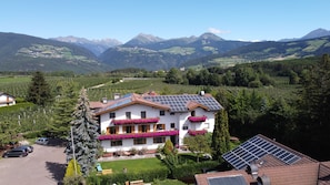 Uma casa de dois andares com painéis solares, rodeada por uma paisagem verdejante e montanhas.