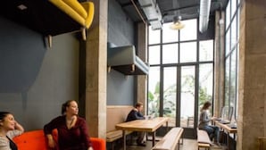 Un espacio interior moderno con techo alto, ventanas amplias y una variedad de zonas de estar, incluyendo un sofá rojo, mesas de madera y bancos.