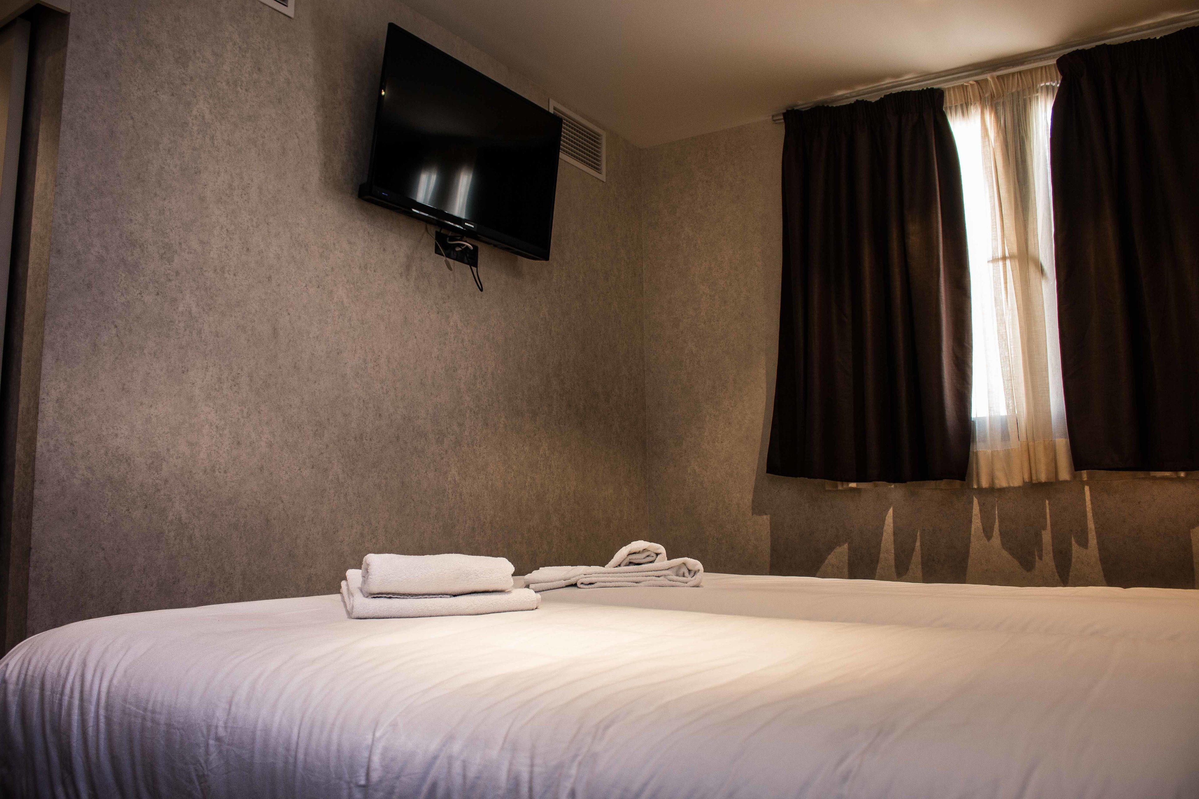 A hotel room with a bed, a flat-screen TV mounted on the wall, and a window with dark curtains.