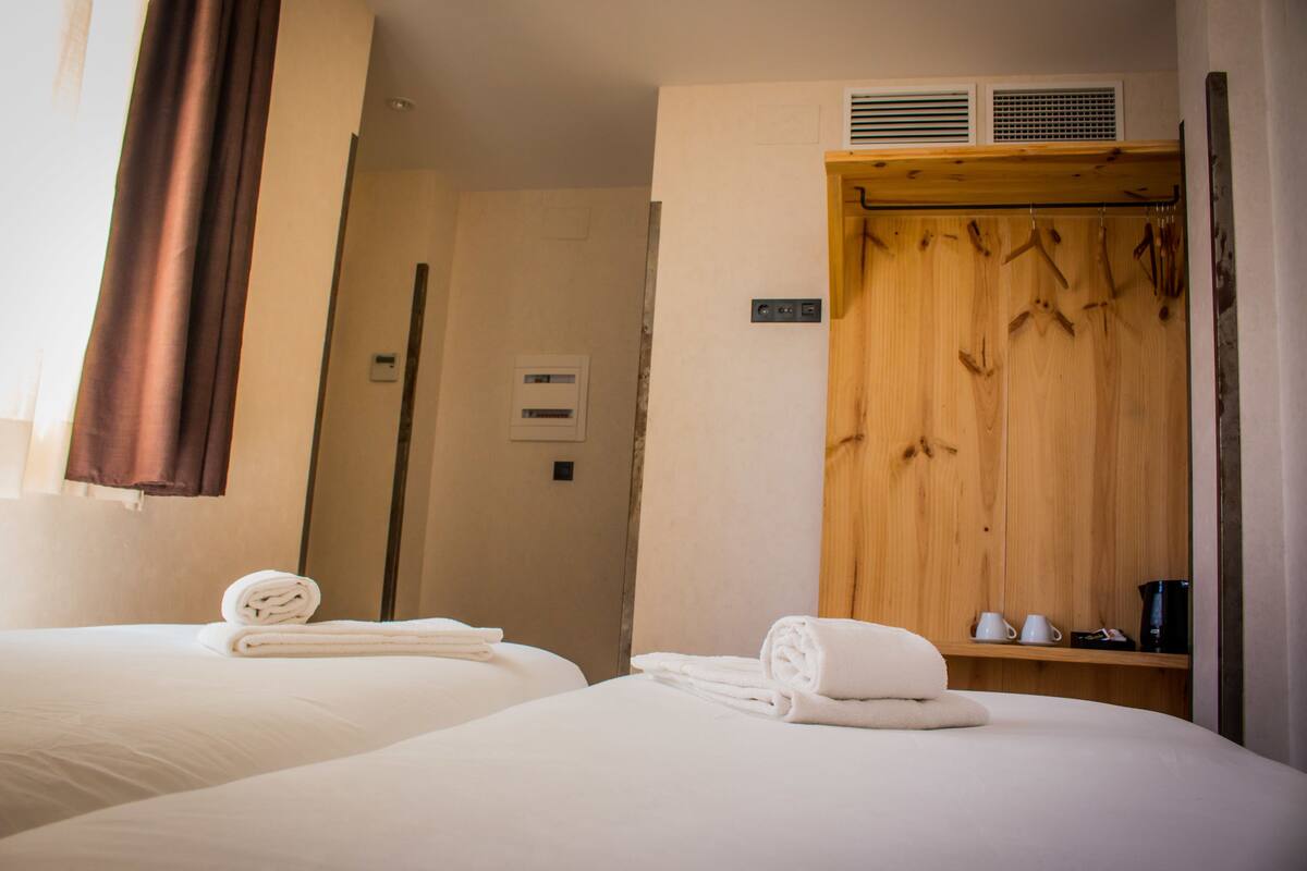 Habitación de hotel con dos camas, un armario de madera, una pequeña estantería con tazas y una cafetera, y una puerta que da a otra habitación.
