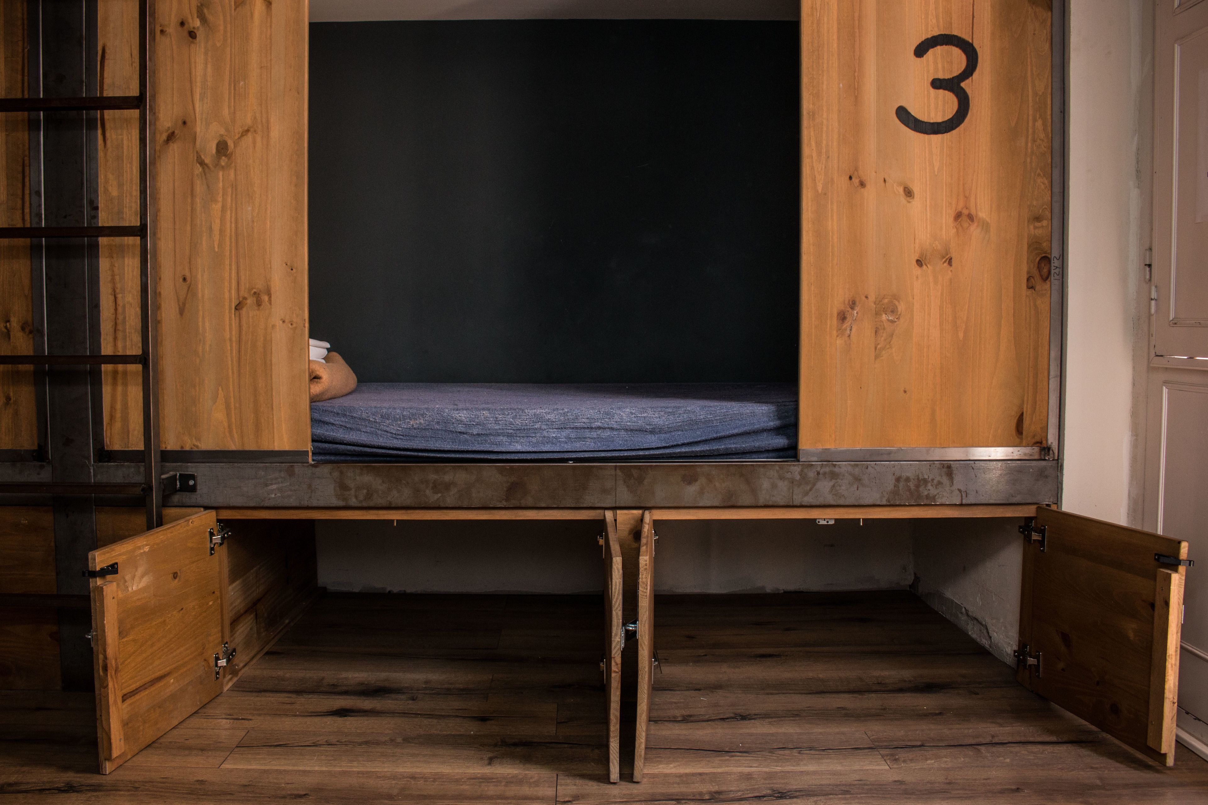 Een houten stapelbed met matras, een nummer 3 op de muur, en een houten ladder.
