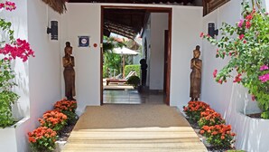 Entrada de hotel con felpudo rojo, dos estatuas de madera y plantas en macetas con flores rojas.
