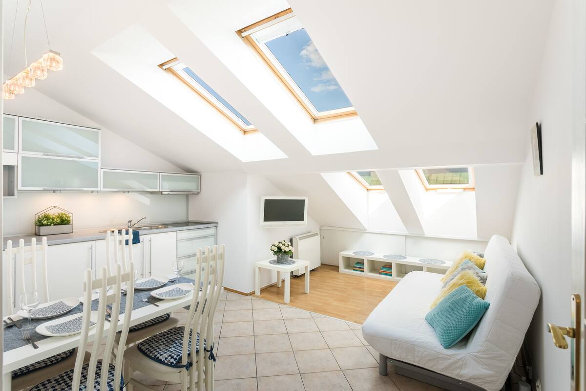 A modern loft apartment with skylights, a dining area, a kitchen, and a living space with a sofa and cushions.