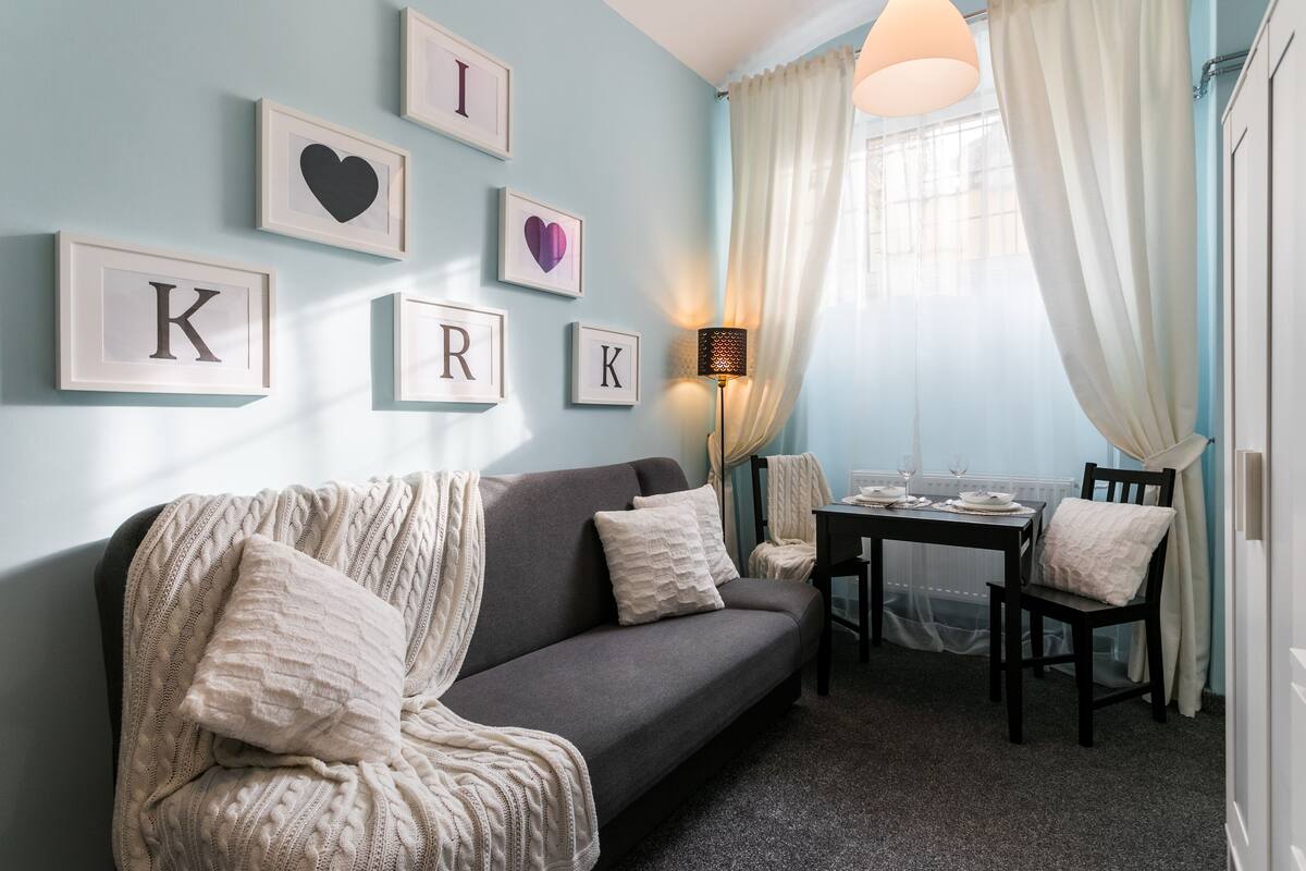 A cozy living room with a grey sofa, a small dining table, and a lamp. There are framed letters and hearts on the wall.