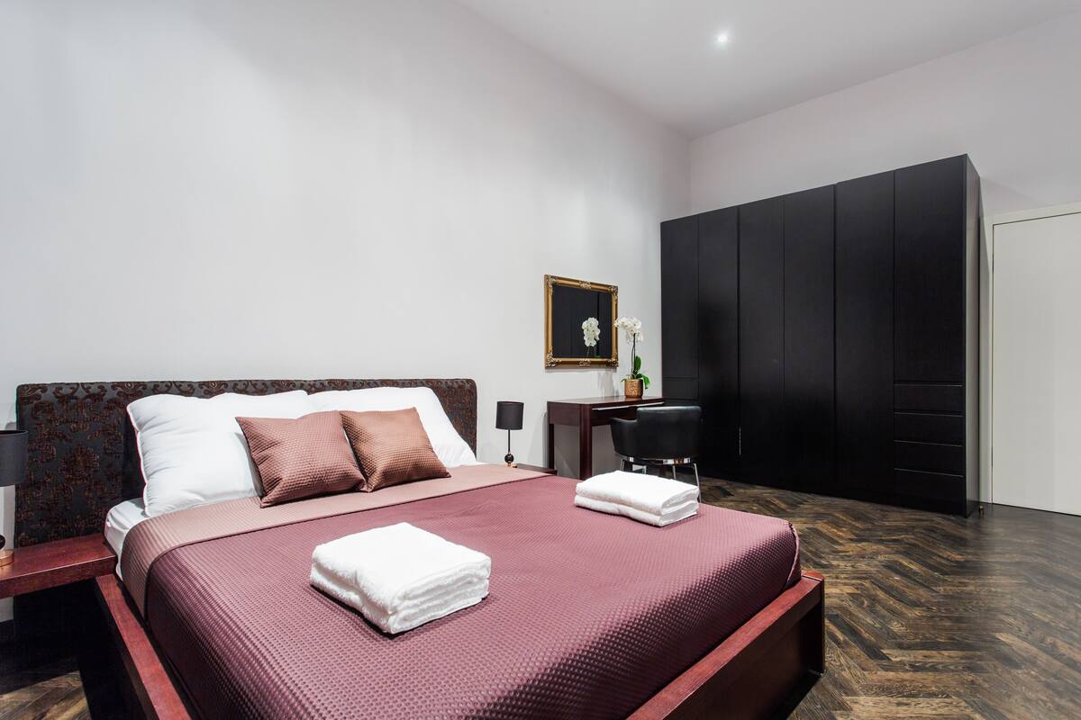 A modern hotel room with a large bed, a desk, and a wardrobe.