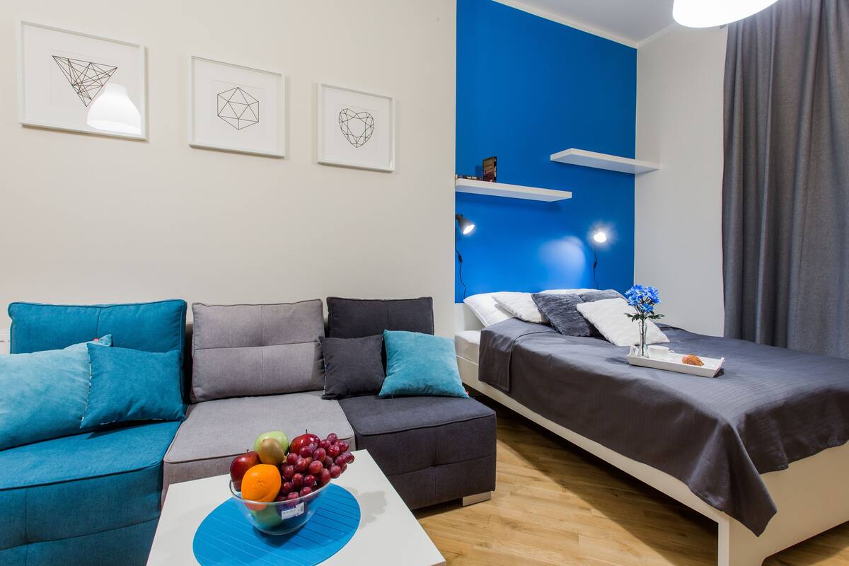A modern room with a sofa, a bed, and a coffee table with fruit. There are wall art pieces and a blue accent wall.