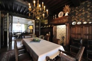 A dining area with a long table set for four, surrounded by wooden chairs. A chandelier hangs from the ceiling, and a fireplace is visible in the background.