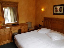 Et hotelværelse med en seng, et skrivebord med skuffer, en stol, en telefon, en lampe og et billede p å væggen.