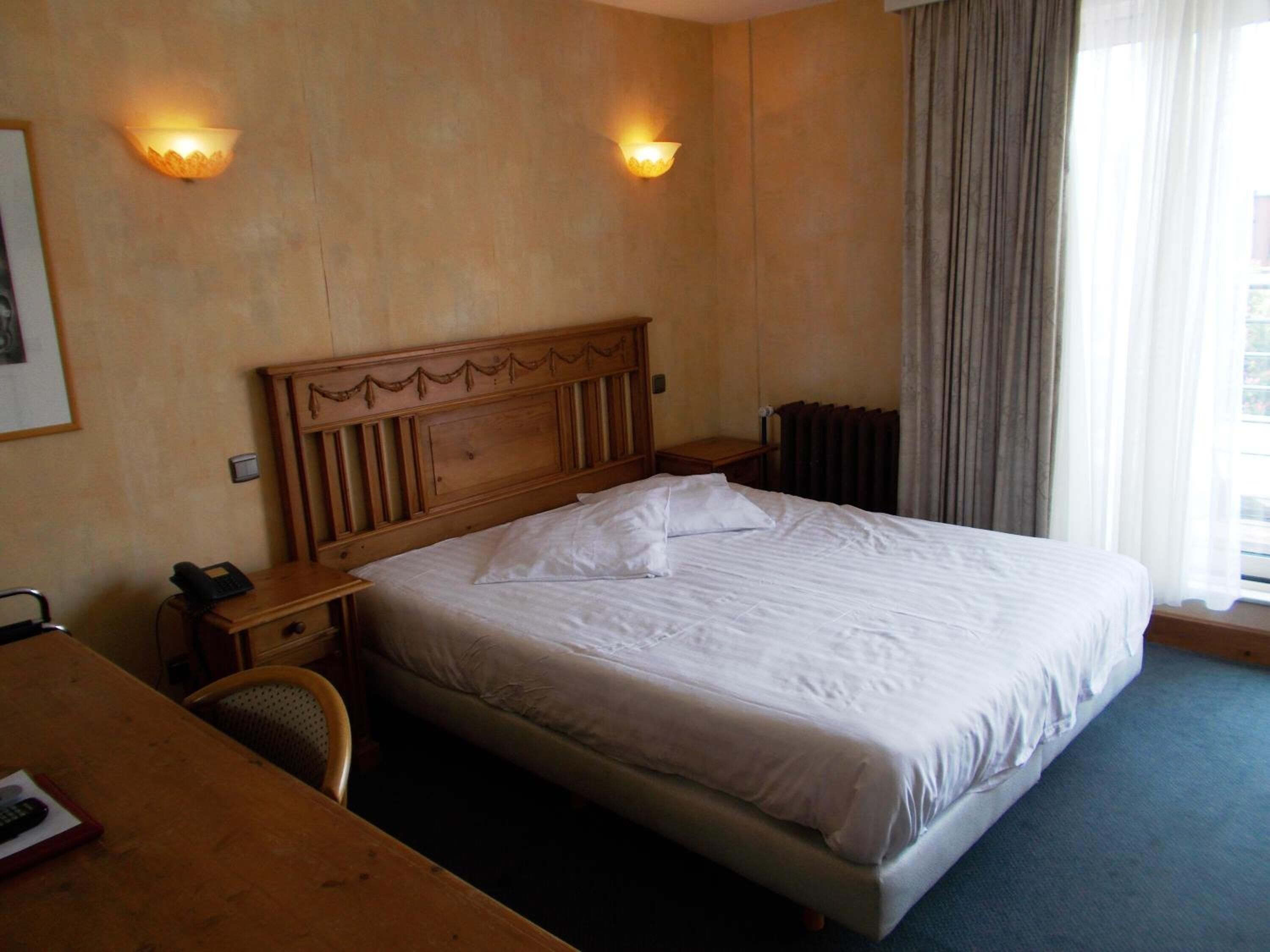 Quarto de hotel com cabeceira de madeira, duas luminárias de apoio, uma escrivaninha com telefone e uma cadeira.